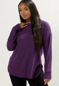 Maglietta a maniche lunghe viola con scollatura rotonda, vestibilità rilassata, spacco laterale e texture morbida, abbinata a pantaloni neri. Nessun motivo o accessorio visibile.