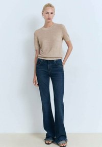 Beige tekstureret striktop med korte ærmer, parret med flare mørkeblå jeans, der har forlommer og diskret kantdetaljering. Sorte sandaler.