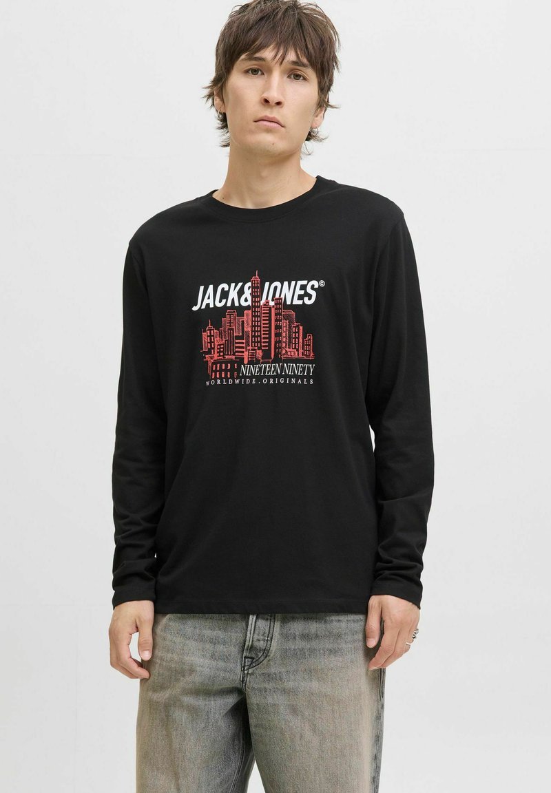 Joven vistiendo una camiseta negra de manga larga con un skyline urbano rojo y el texto "JACK & JONES NINETEEN NINETY", combinada con unos jeans grises.