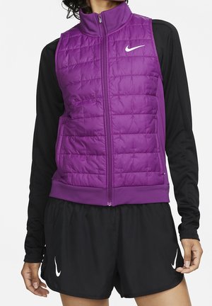 Violetti tikattu Nike-liivi vetoketjulla edessä, päällä musta pitkähihaisen paita ja mustat Nike-urheilushortsit seisovalla henkilöllä.