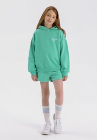 Sudadera con capucha verde menta y pantalones cortos a juego. Texto del logo en blanco en el pecho. El modelo lleva zapatillas blancas y calcetines hasta la rodilla con rayas azul claro.