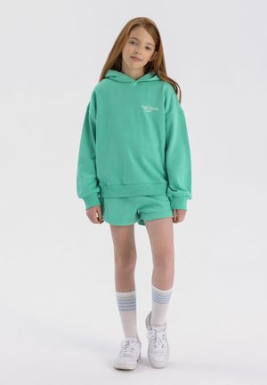 Felpa con cappuccio verde menta abbinata a pantaloni corti. Testo del logo bianco sul petto. Il modello indossa sneaker bianche e calze fino al ginocchio con strisce azzurre.