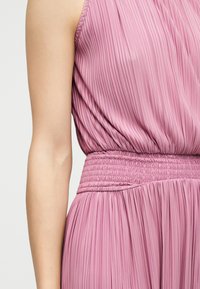 Ροζ αμάνικο πλισέ φόρεμα με ελαστική μέση, που δείχνει τον άνω κορμό και το αριστερό χέρι σε ουδέτερο φόντο.