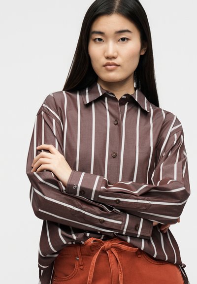 Γυναίκα με μακριά σκουρόχρωμα μαλλιά φοράει πουκάμισο με κάθετες ρίγες σε καφέ και λευκό χρώμα, κουμπωμένο, και παντελόνι σε σκουρόχρωμο πορτοκαλί με ζώνη δεμένη, με σταυρωμένα χέρια.