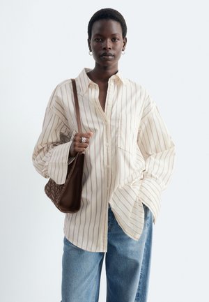 Femme aux cheveux courts portant une chemise rayée oversize et un jean bleu, tenant un sac à main tissé marron sur l'épaule.