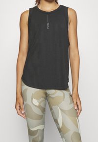 Czarny prążkowany top na ramiączkach z okrągłym dekoltem i subtelnym białym logo. W połączeniu z beżowymi legginsami w kamuflażowy wzór. Gładka faktura materiału.