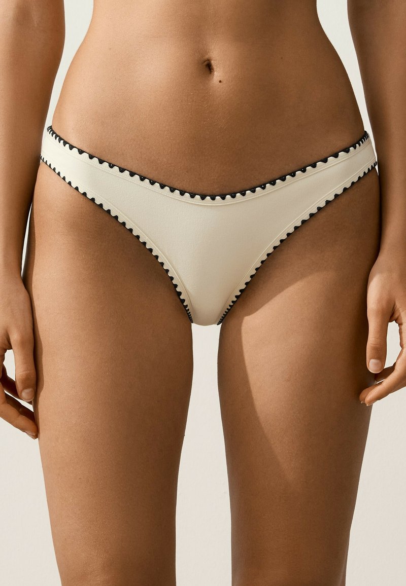 Partie médiane d'une femme portant un bas de bikini blanc avec un bord festonné noir sur un fond neutre.