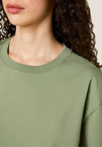 Olīvu zaļš sporta krekls ar apaļu kakla izgriezumu, ar ribotu maliņu un gludu tekstūru. Audums izskatās mīksts un ikdienišķs, ar brīvu piegriezumu.