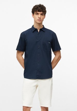Jeune homme aux cheveux bouclés portant une chemise bleu marine à manches courtes boutonnée et un short blanc, debout les mains dans le dos.