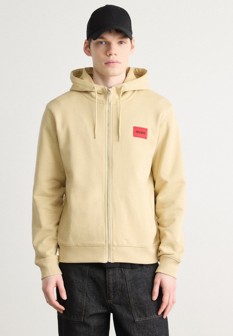 Hooded krekls gaisā bēšā krāsā, ar priekšējo rāvējslēdzēju un sarkanu HUGO logotipa uzlīmi. Kopā ar tumšām, brīvām biksēm.