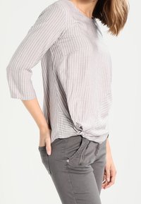 Gestreifte graue Bluse mit Dreiviertelärmeln, vorn mit Knoten-Detail, aus leichtem Stoff. Kombiniert mit grauen, engen Hosen mit Reißverschlüssen.