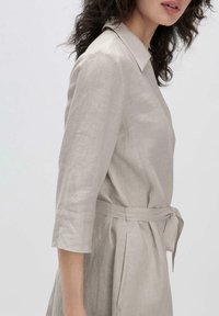 Abito in lino beige chiaro con collo a camicia, maniche a tre quarti, vita con fiocco e tasche laterali. Tessuto morbido e vestibilità rilassata.