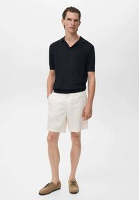 Camisa polo negra de manga corta combinada con pantalones cortos blancos y zapatos de cordones marrón claro, mostrando una textura suave y un ajuste relajado.