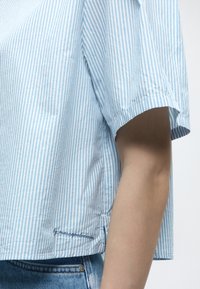 Chemise à manches courtes à rayures verticales bleu clair et blanc, portée avec un jean en denim bleu, montrant la couture latérale et le bras.
