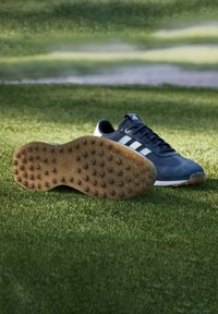 Scarpe da ginnastica blu con tre strisce bianche, finitura in suede testurizzato, suola in gomma con tacchetti e linguetta imbottita, posate su erba sintetica verde.