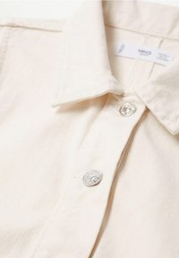 Chemise beige à col avec boutons-pression argentés et étiquette de marque visible à l'intérieur du col.