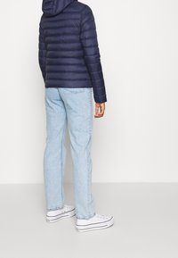 Chaqueta de plumas azul marino con costuras horizontales, capucha, combinada con jeans azul claro y zapatillas blancas. Enfoque en la vista posterior.
