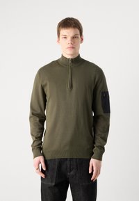 Dunkelgrüner Reißverschluss-Pullover mit hohem Kragen, gerippten Bündchen und einem schwarzen Stoffpatch am linken Arm. Glatter Strickstoff.