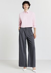 Frau in einem hellrosa, kurzen, knöpfbaren Hemd, hochtaillierten grauen gestreiften, weiten Hosen und weißen spitzen Absatzschuhen, die aufrecht steht.