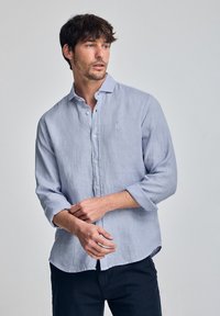 Jasnoniebieska lniana koszula z kołnierzykiem zapinanym na guziki, długimi rękawami podwiniętymi, z małym haftowanym logo na piersi.