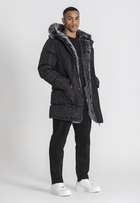 Sort vinterjakke med faux fur-fôret hætte, der har lynlås og knaplukning, iført over en mørk skjorte og sorte jeans, med hvide sneakers.