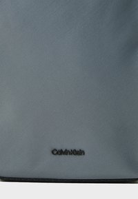 Šedá texturovaná tkanina s vertikálními pruhy, na spodní části s černým logem "Calvin Klein". Ozdobena tmavým lemováním.
