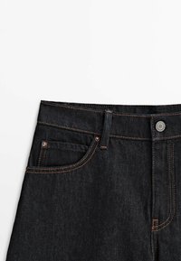 Mörk denimjeans med hög midja, som har orange sömmar, en framficka och en metallknappslutning vid midjan.