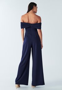 Marineblauwe off-the-shoulder jumpsuit met een getailleerde taille en wijd uitlopende broekspijpen. Heeft een gerimpelde buste en een verborgen rits aan de achterkant.
