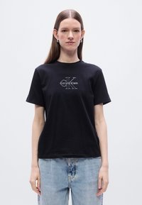 Μαύρο βαμβακερό t-shirt με λευκό λογότυπο "Calvin Klein" και περίγραμμα "CK". Κοντά μανίκια και στρογγυλή λαιμόκοψη.