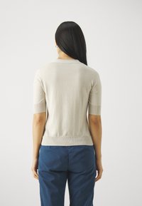 Beżowy sweter z krótkim rękawem, z ribbowanym wykończeniem u dołu i przy dekolcie, prezentujący dopasowany krój i miękką teksturę z tyłu. W połączeniu z niebieskimi spodniami.