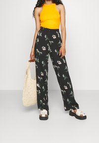 Kobieta ubrana w pomarańczowy top bez rękawów i czarne, kwiatowe spodnie szerokie, trzymająca plecioną beżową torbę na ramię z drewnianymi uchwytami.