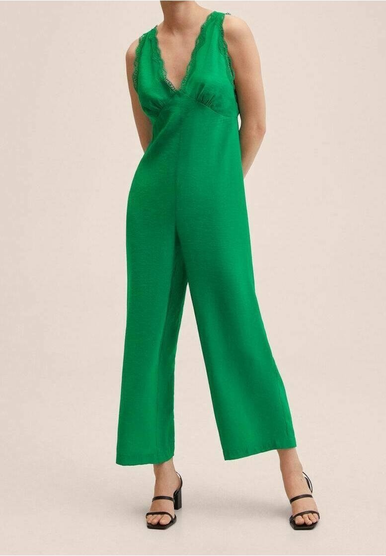 Femme portant une combinaison verte sans manches avec bordure en dentelle et jambes larges, associée à des sandales noires à talons et à brides.
