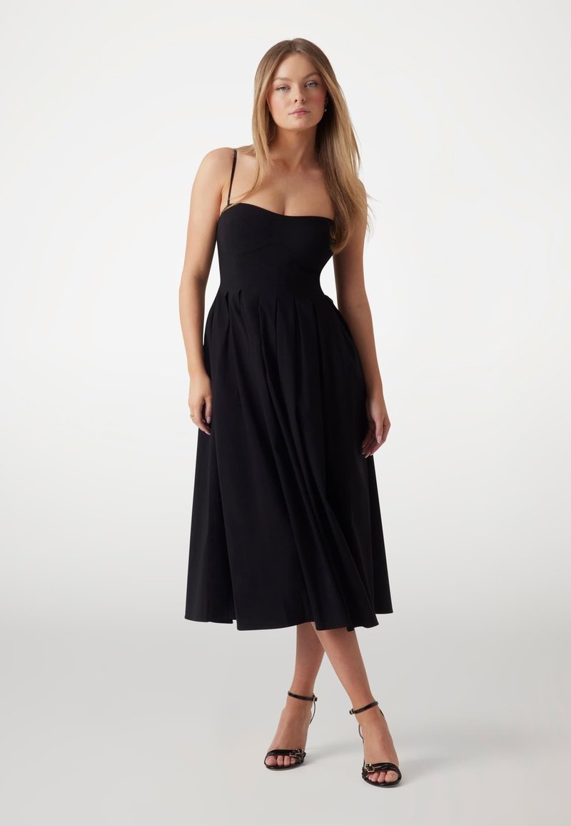 Robe noire, longueur genoux, avec bretelles spaghetti, corsage ajusté et jupe plissée. Tissu lisse avec une finition mate.