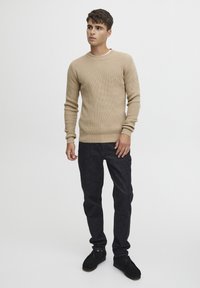 Beige textuurtrui met een ronde hals, gecombineerd met donkere denim jeans en zwarte schoenen. De stof vertoont een geribbelde structuur.