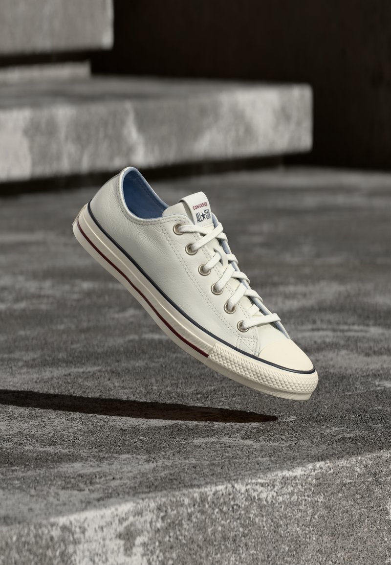 Λευκό δερμάτινο sneaker Converse All Star χαμηλού προφίλ που αιωρείται πάνω από μια γκρι τσιμεντένια επιφάνεια με πέτρινα σκαλιά στο φόντο.