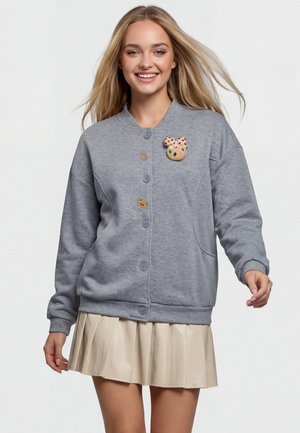 Ung kvinde med langt blondt hår, smilende, iført en grå cardigan med knapper og legesyge pins samt en beige plisseret nederdel.