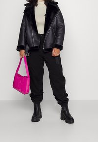 Giacca biker in finta pelle nera con colletto in pelliccia, dolcevita crema, pantaloni neri a gamba stretta e borsa lucida rosa. Stivali con tacco spesso.