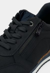 Sneaker noir avec des lacets épais, une fermeture éclair sur le côté, une texture perforée et une semelle bleue et blanche avec un accent en liège.