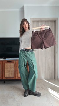 Asmuo laiko rudos spalvos kordurovinį šortą ant medinio kablio, dėvi žalias kargo kelnes ir pilką megztą megztinį, stovi kambaryje.