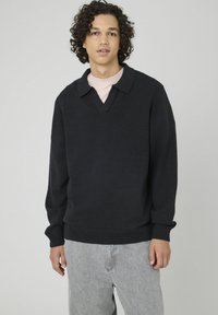 Jeune homme aux cheveux bouclés portant un pull noir avec col sur une chemise claire et un pantalon gris clair, se tenant devant un fond uni.