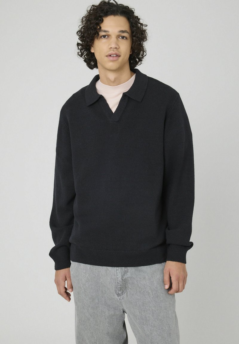 Jeune homme aux cheveux bouclés portant un pull noir avec col sur une chemise claire et un pantalon gris clair, se tenant devant un fond uni.