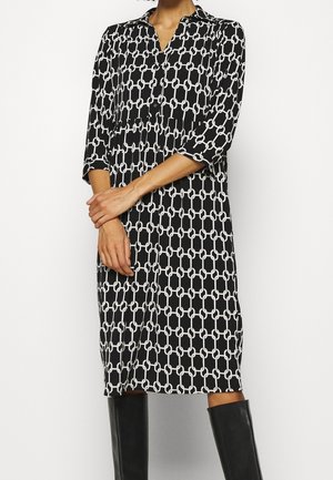 Kvinde iført en sort knælængde kjole med hvidt kædemønster og trekvart-ærmer, kombineret med sorte knæhøje støvler.