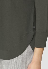 Dunkelgrüner, gerippter Pullover mit abgerundetem Saum und ärmellangen Ärmeln, kombiniert mit einem hellgrauen Textilstoff darunter. Kleines Logetikett sichtbar.