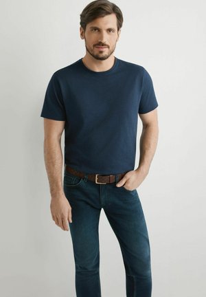 Mężczyzna z brodą ubrany w granatowy t-shirt, ciemne dżinsy i brązowy pleciony pasek stoi z jedną ręką w kieszeni.