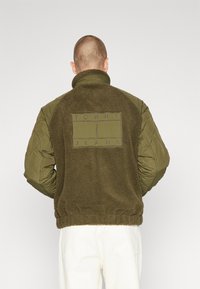 Chaqueta verde oliva con un cuerpo de forro polar texturizado y mangas acolchadas. Presenta un logotipo rectangular en la parte de atrás con el texto "TOMMY JEANS".