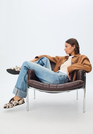 Giacca oversized marrone, top bianco, jeans azzurri chiari, scarpe a fantasia. Seduto su una sedia in pelle marrone con gambe in cromo. Sfondo semplice.