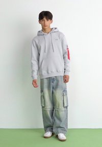 Felpa grigia con tasca a marsupio e lacci, abbinata a pantaloni cargo in denim oversize e sfilacciati con tasche laterali. Sneakers bianche.