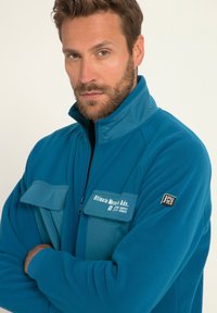 Giacca in pile blu con colletto a zip, dotata di due tasche piatte e una toppa con logo sulla manica. Tessuto morbido e vestibilità rilassata.