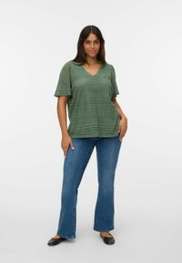 Femme avec de longs cheveux noirs portant un haut à motifs en dentelle vert à manches courtes, un jean flare bleu et des ballerines noires se tenant sur un fond blanc.
