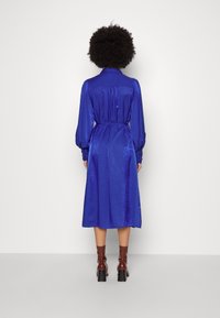 Blå sidentklänning med långa puffärmar och en formsydd midja, når halvvägs ner till vaden; har en krage och en slät, reflekterande yta.